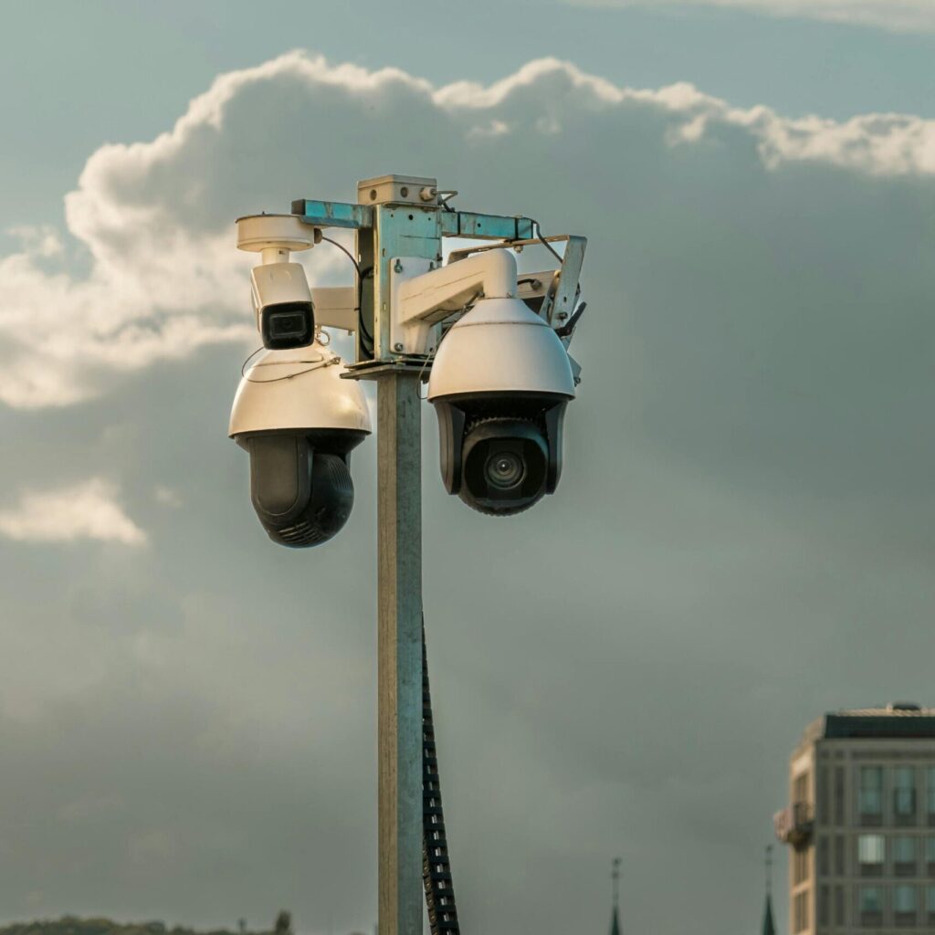 Two modern surveillance cameras mounted on a pole in an urban setting under a cloudy sky.